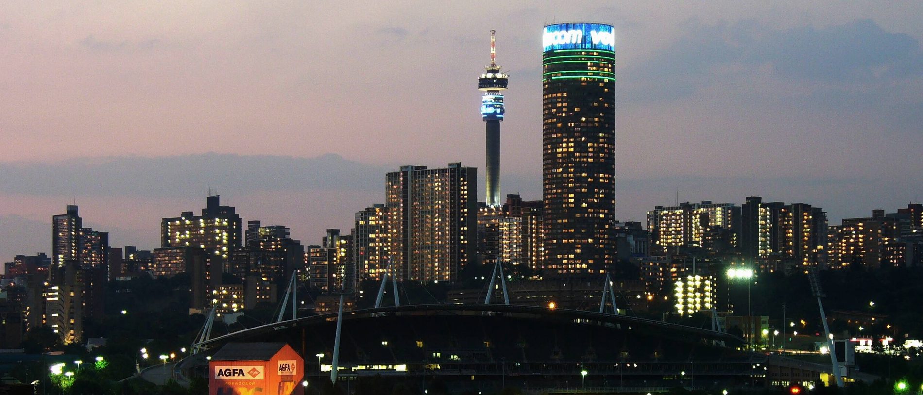 The Johannesburg skyline at dusk: the Hillbrow telecommunications tower and a cluster of lit high-rise buildings, seen across the city.